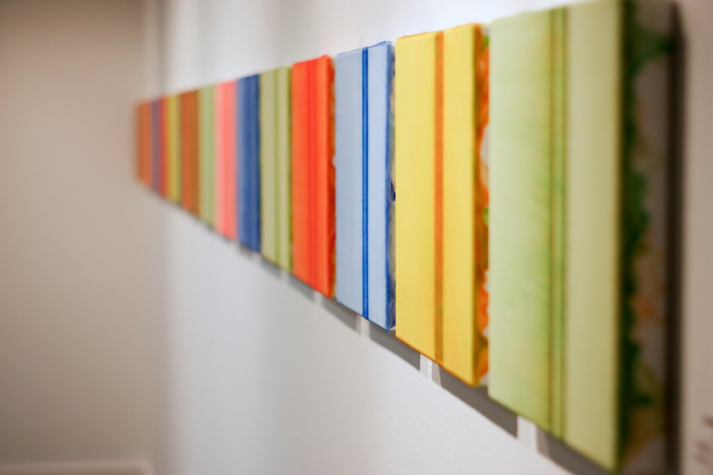 Line of colorful13" square paintings of a line installed in a row.