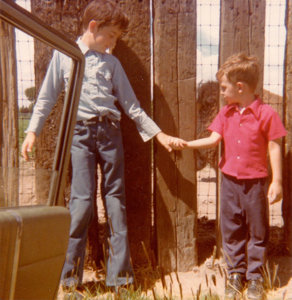 Three year old boy stands in front of a fence holding the hand of his nine year old brother.