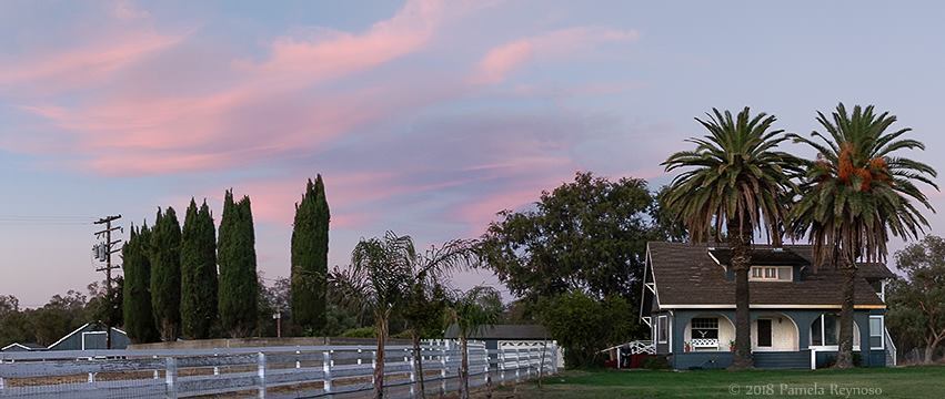 A two story circa 1890's ranch house is shown at the end of a long driveway lined with white fencing at sunset.