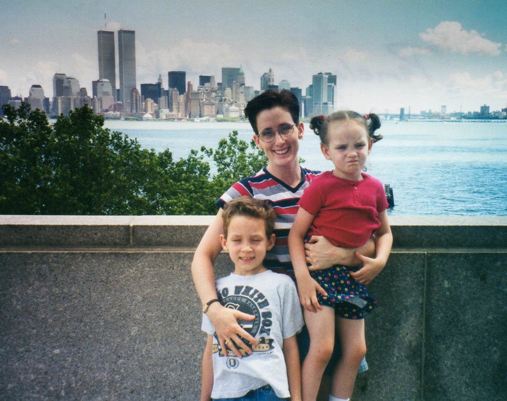 A woman in a red, white, and blue shirt stands on Liberty Island with her 5 yo son and holds her 2yo daughter. The pre-9/11 twin towers are visible across the water.