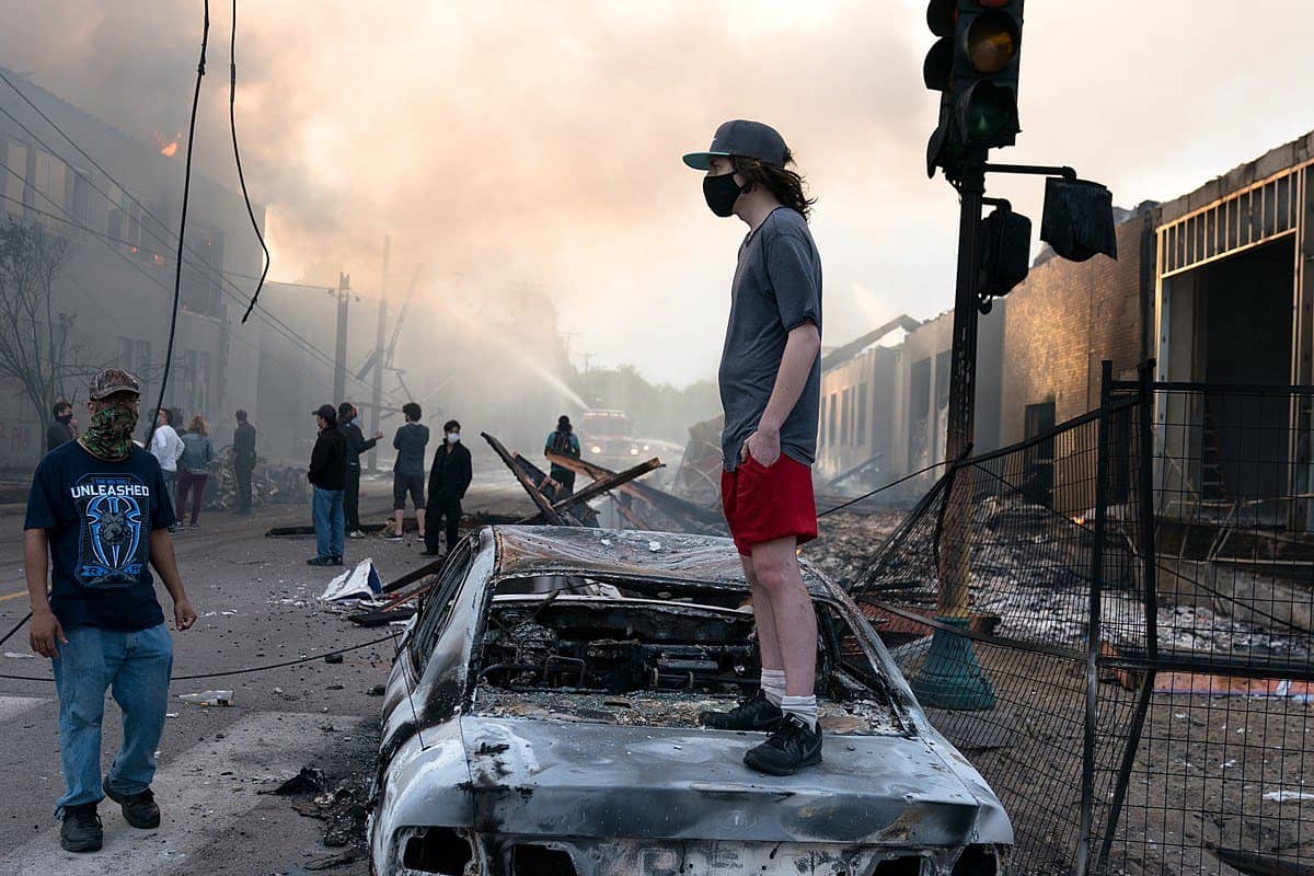 A man stands on the hood of a burned out car in the middle of a street.