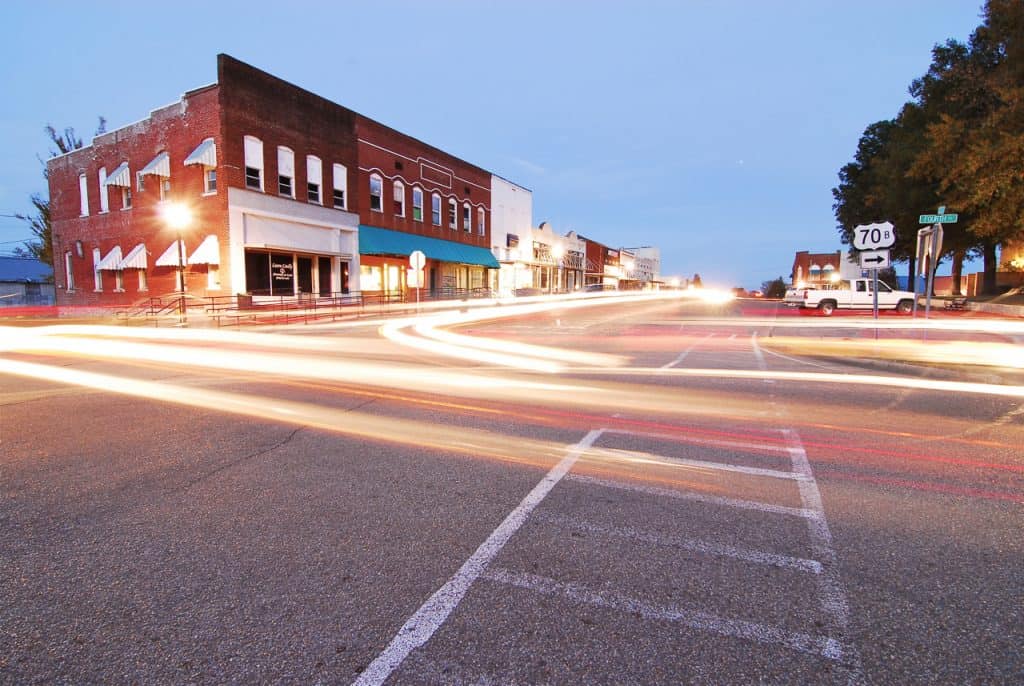 In this dusk long exposure image taken in De Queen, AK, several old buildings with brick facades sit on the side of a street with streaked headlights of cars on the roads.