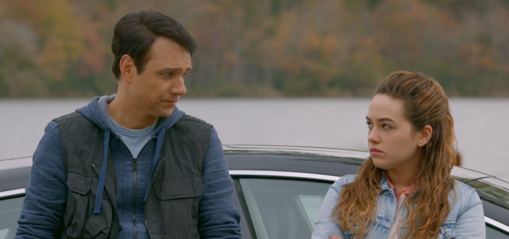 Ralph Macchio and Mary Mouser from The series Cobra Kai stand talking by a car in a screen capture.