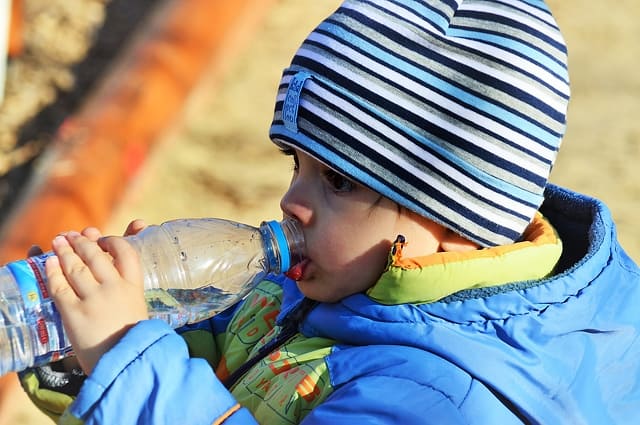 A toddler in a colorful knit hat and blue coat drinks from a water bottle.