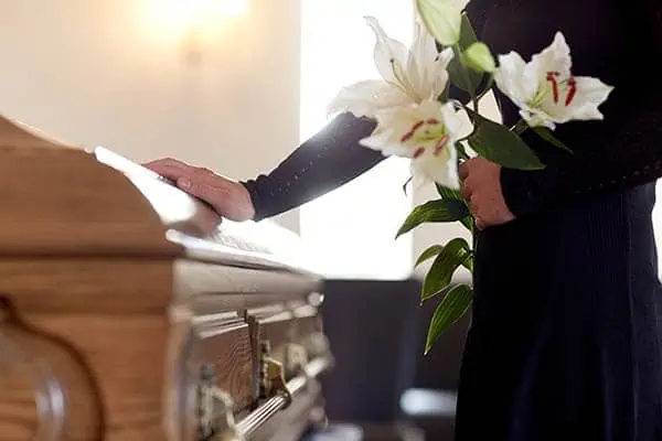 A woman dressed in black is shown with a hand on a casket and holding a bouquet of white flowers.