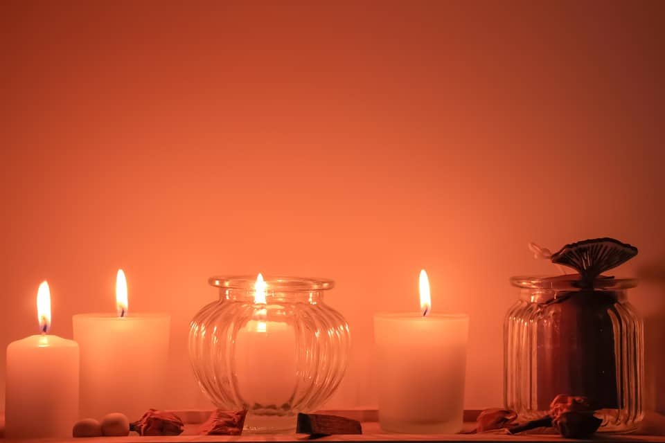 Four short white candles, one in a glass votive, are lit with a soft orange background and whole spices in the foreground.