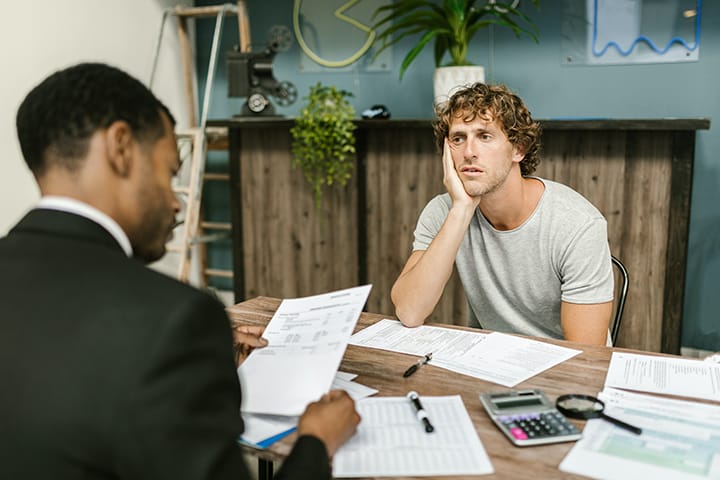 Two men sit across a desk from each other, one in a suit looking at papers, the other in a t-shirt looking overwhelmed.