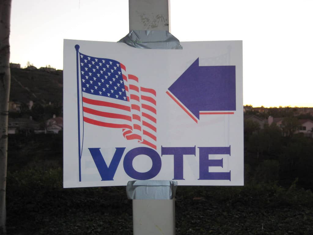 A white sign with a blue arrow pointed toward a United States flag with the word Vote below them is taped to a pole with duct tape.