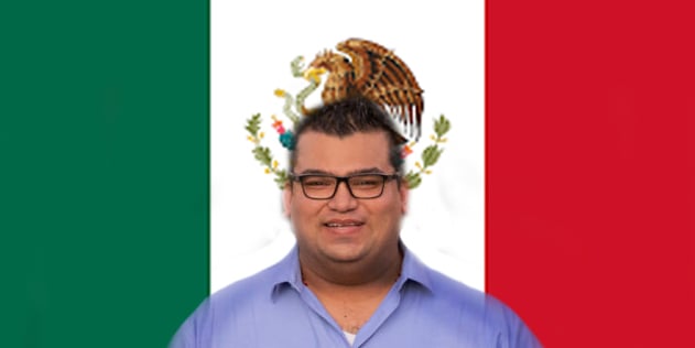 A man with glasses and dressed in a blue shirt stands in front of the flag of Mexico.
