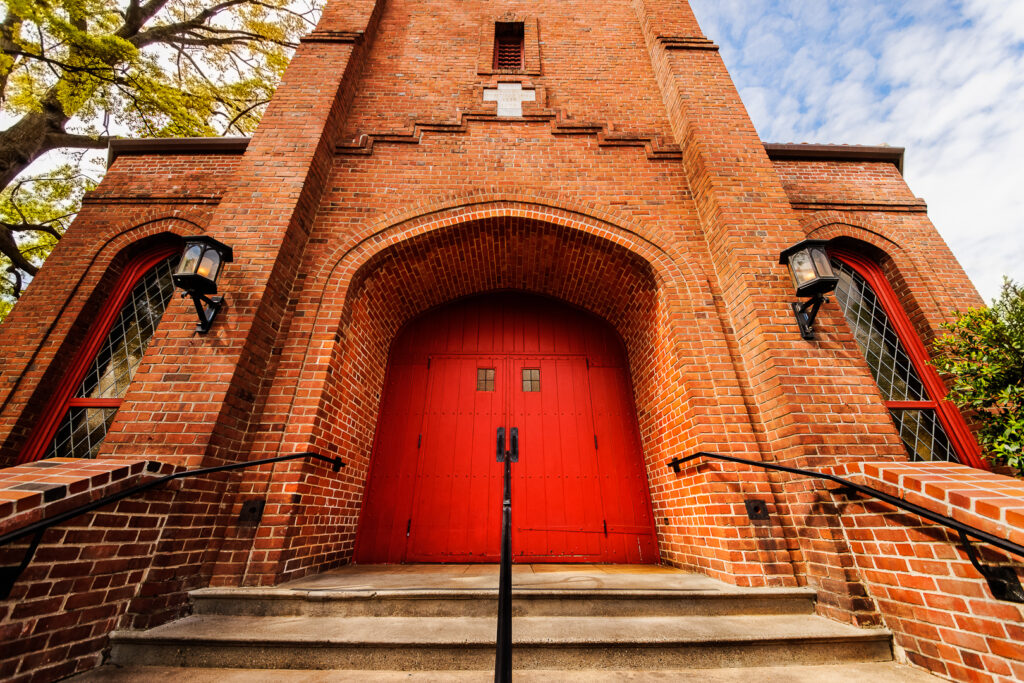 An old red brick church building with red double doors, a white stone cross above them and matching leaded clear glass windows sits at the top of cement stairs in Chico, California, photographed by Pamela Reynoso