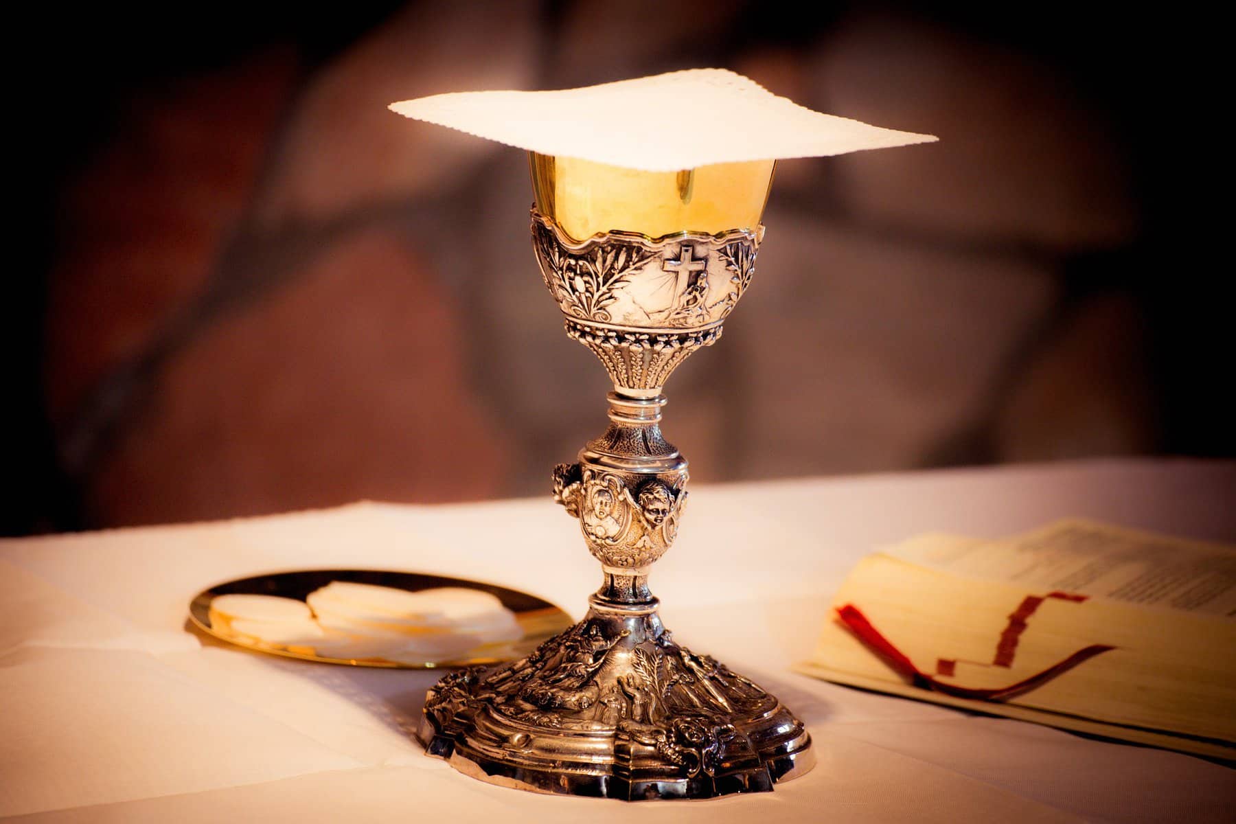 A silver and gold engraved communion chalice sits on an altar with communion wafers sitting behind it on a small plate.