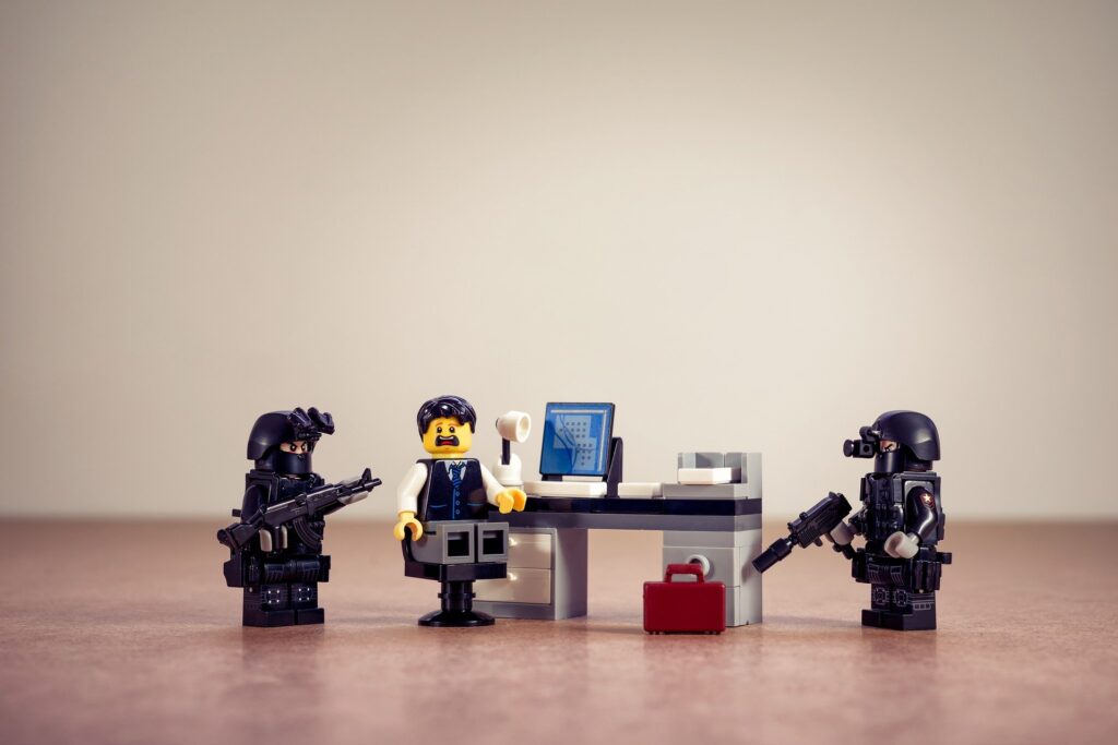 A Lego minifig sits in a chair at a desk with a surprised look. To each side is a minifig dressed in police-type gear with long guns.