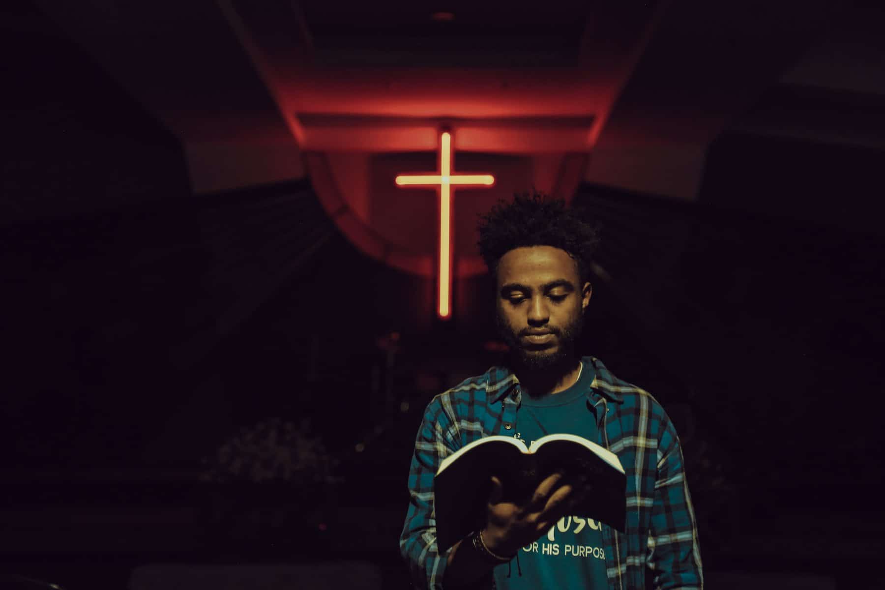A Black man wearing a flannel shirt over a t-shirt holds a softcover bible open in front of him with a neon orange cross placed up high behind him.