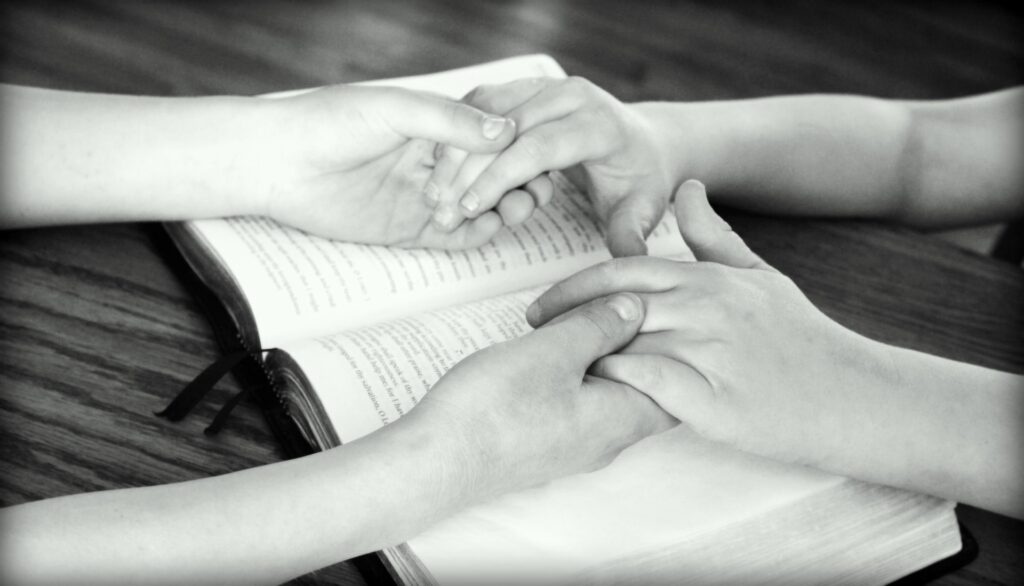 Two children hold hands that are resting on an open bible placed on a table top.