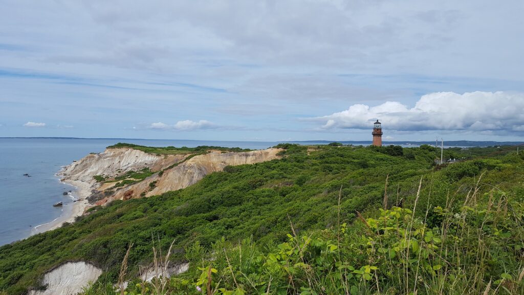 A lighthouse sits in the distance on a sandy-looking cliff surrounded by vegetation and the ocean.
