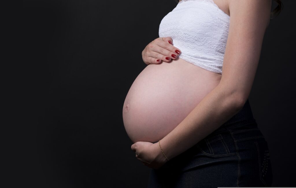 A heavily pregnant woman in jeans and a white tube top is only seen from the hips to the neck holds her bare belly in a studio photo session