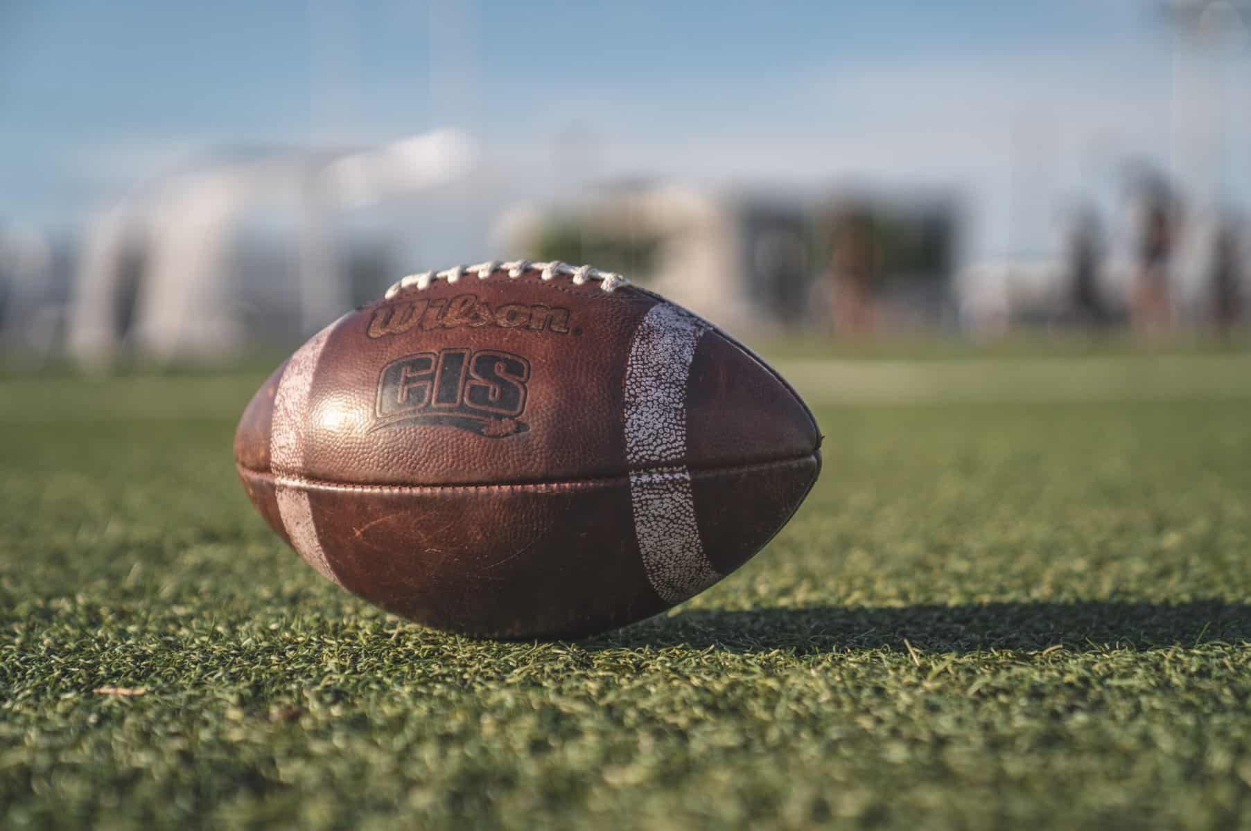 A worn Wilson brand football sits in daylight on an astroturf field and is photographed from close up with the background very blurry