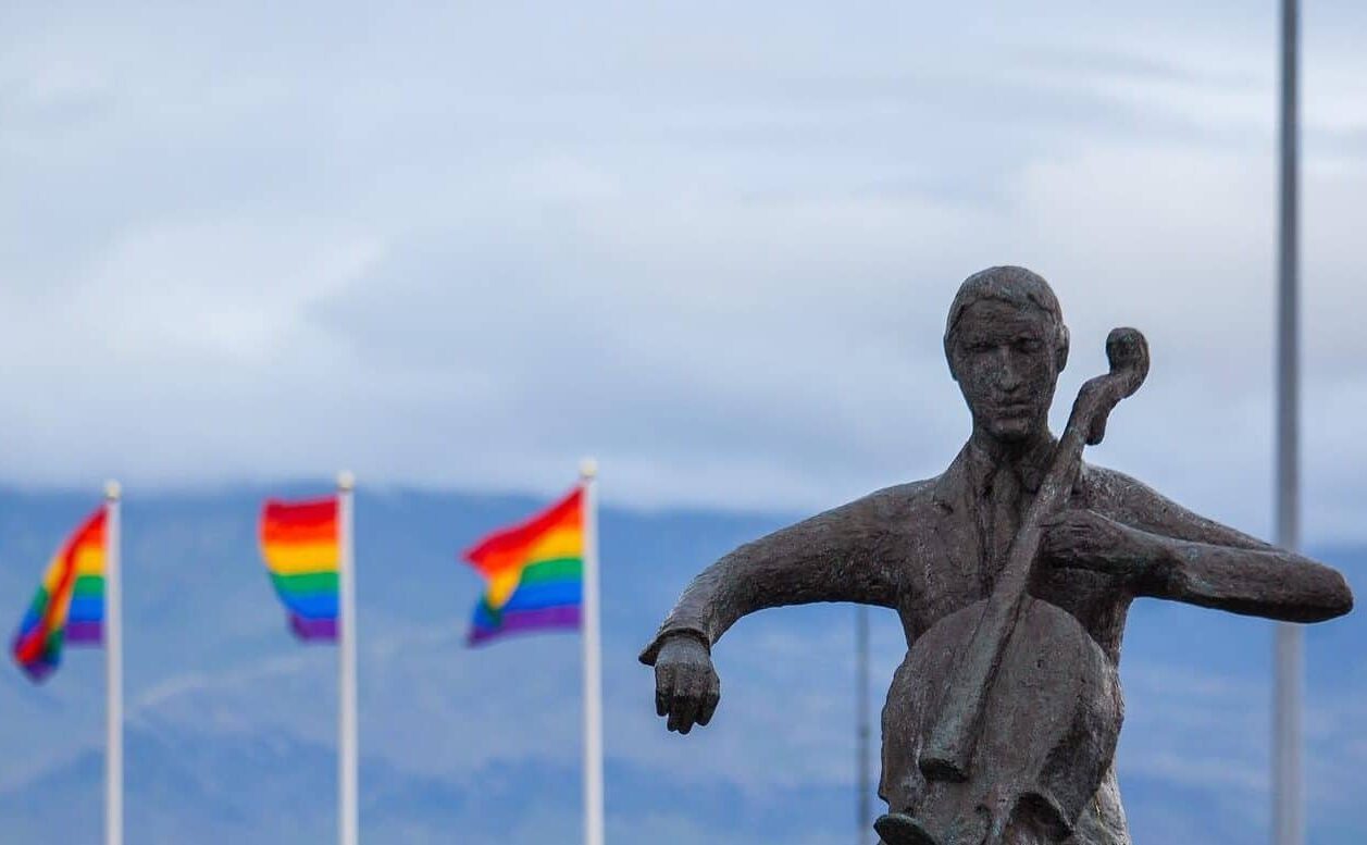 a rough statue of a violin player is seen from the hips up and in the background are three rainbow flags
