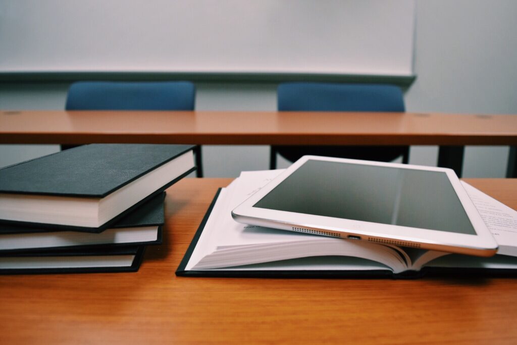 A tablet sits on an open book next to a stack of books on a classroom table.