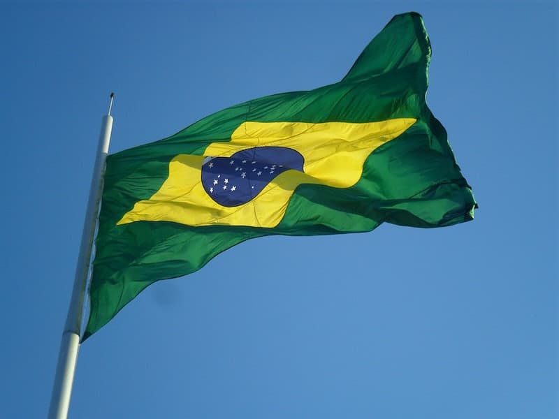 The flag of Brazil is shown at the top of a flag pole waving in the wind against a pure blue sky