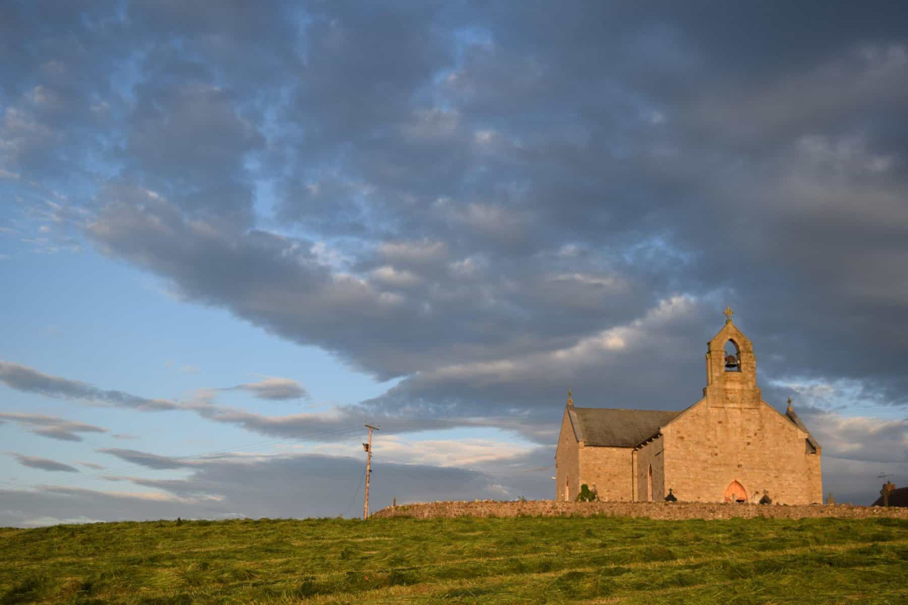 a stone church sits in a field with golden light and shining on it, a blue sky with gray clouds