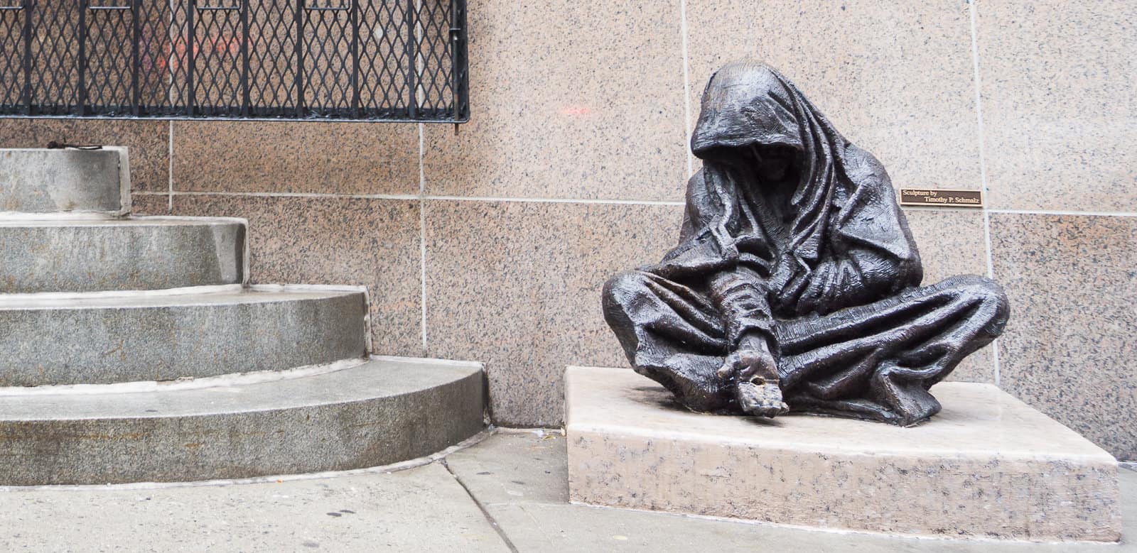 A bronze outdoor sculpture of a begging woman with her hand extended while wrapped in a blanket.