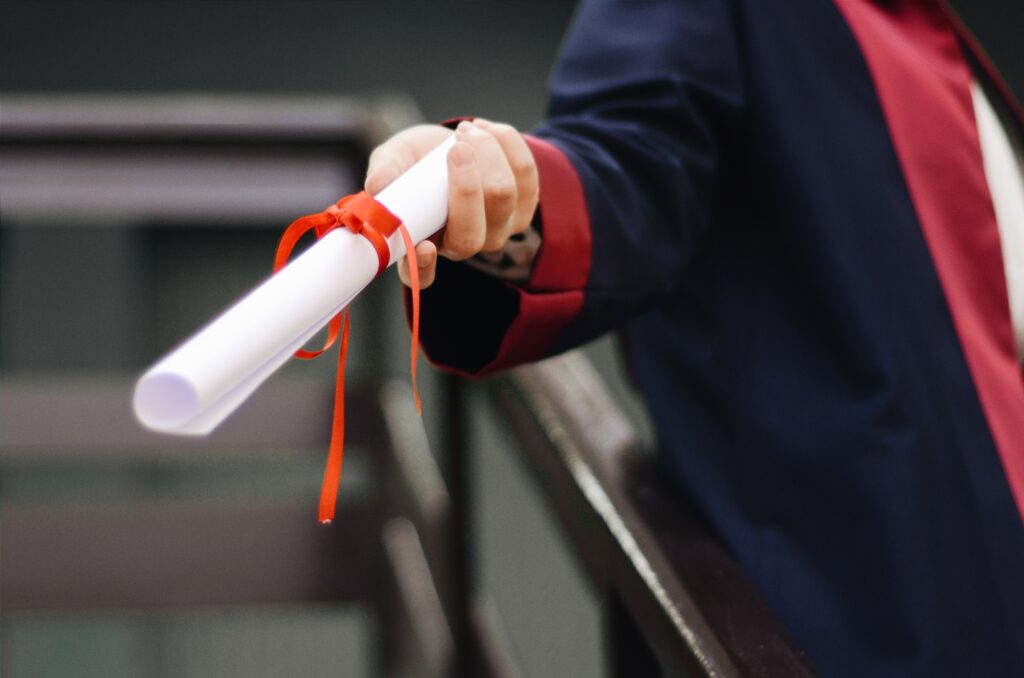 a hand in a navy blue and red academic robe is extended holding a rolled white paper tied with a red ribbon