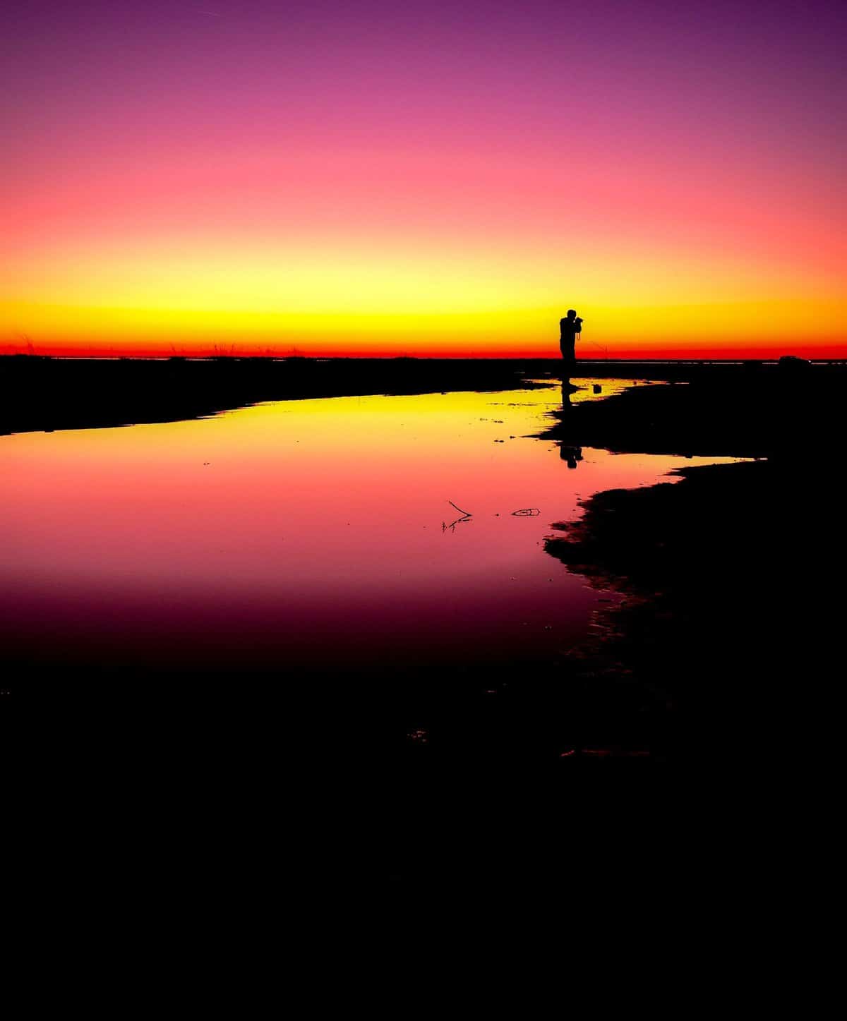 a person stands in silhouette holding a camera against a sunset surrounded by low water as if in a marsh