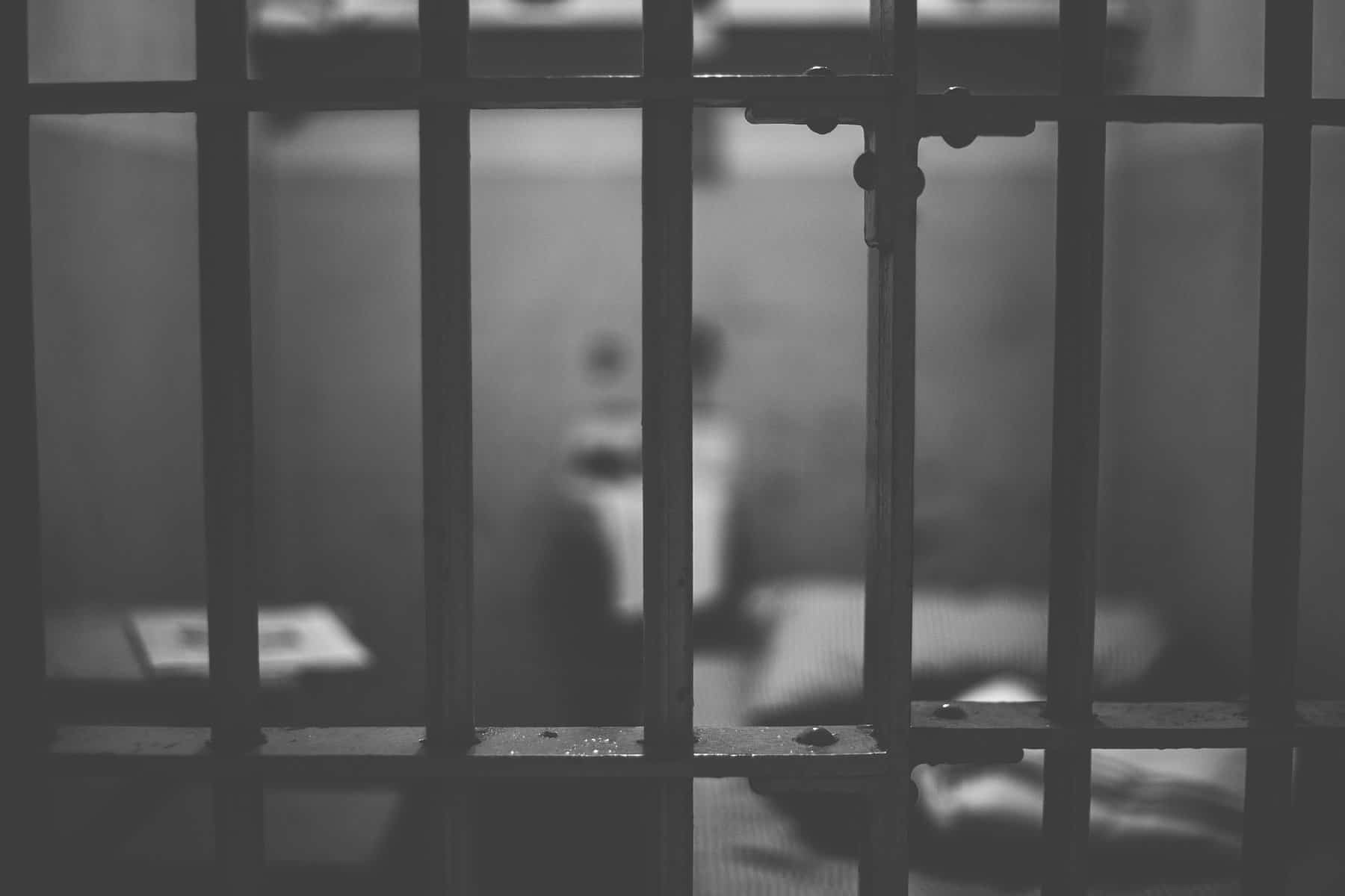 a black-and-white image of in-focus prison bars with a jail cell out of focus behind