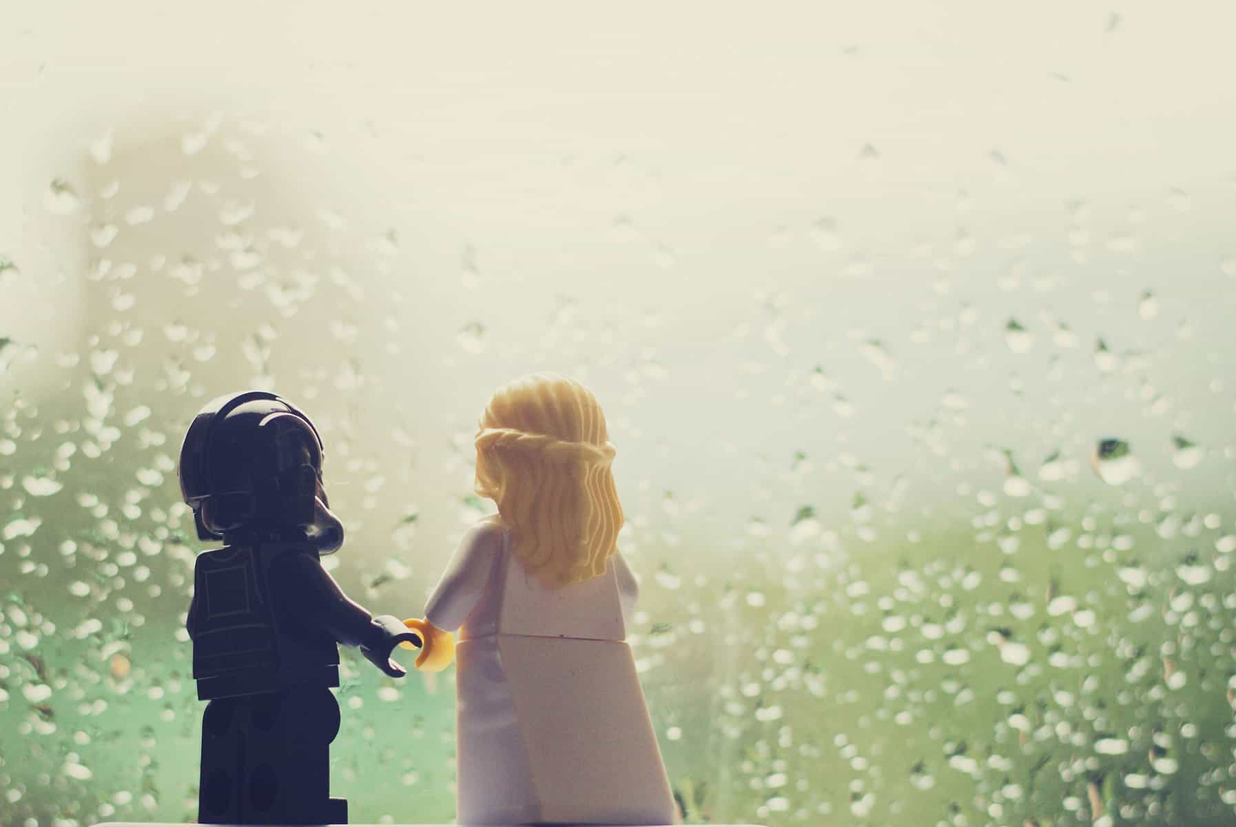 two lego figurines, a space person and a girl with golden hair are seen from the back with a rain-covered window in front of them
