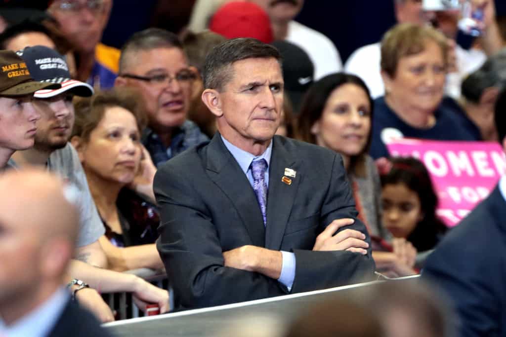 Michael Flynn stands in a suit with arms crossed and looking forward with a crowd behind him