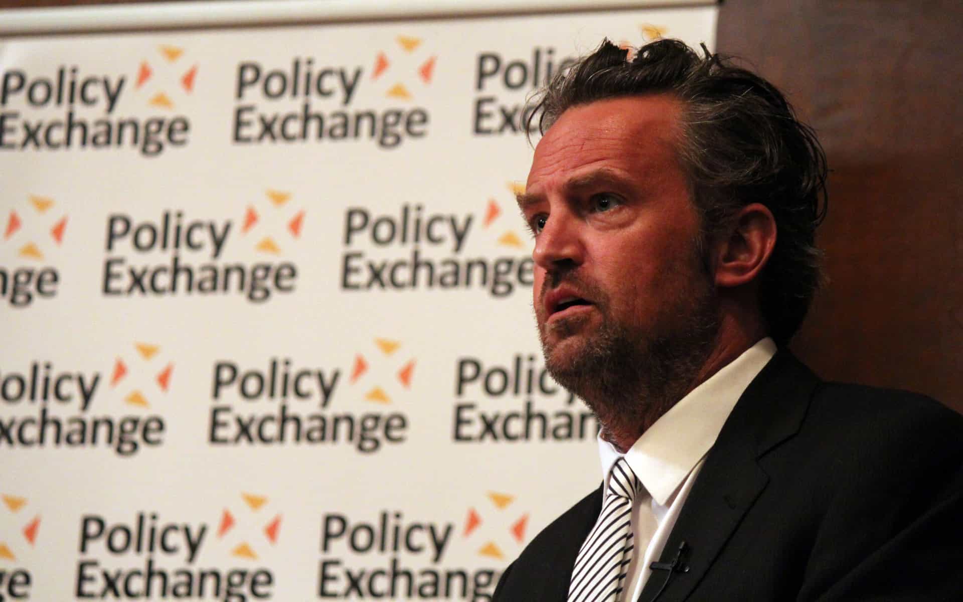 Matthew Perry, in a suit and tie, stands in profile view in front of a branded background