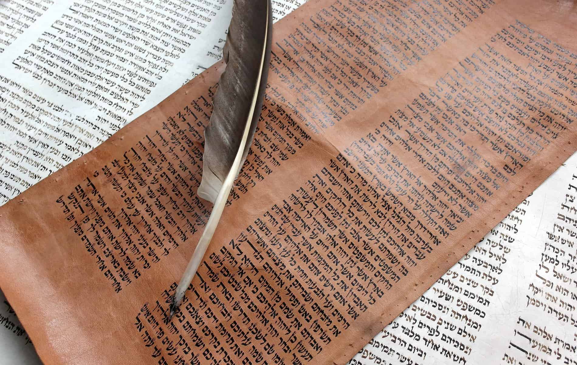 A very old looking dark piece of parchment with a feather quill lays over a whitepaper with words on.