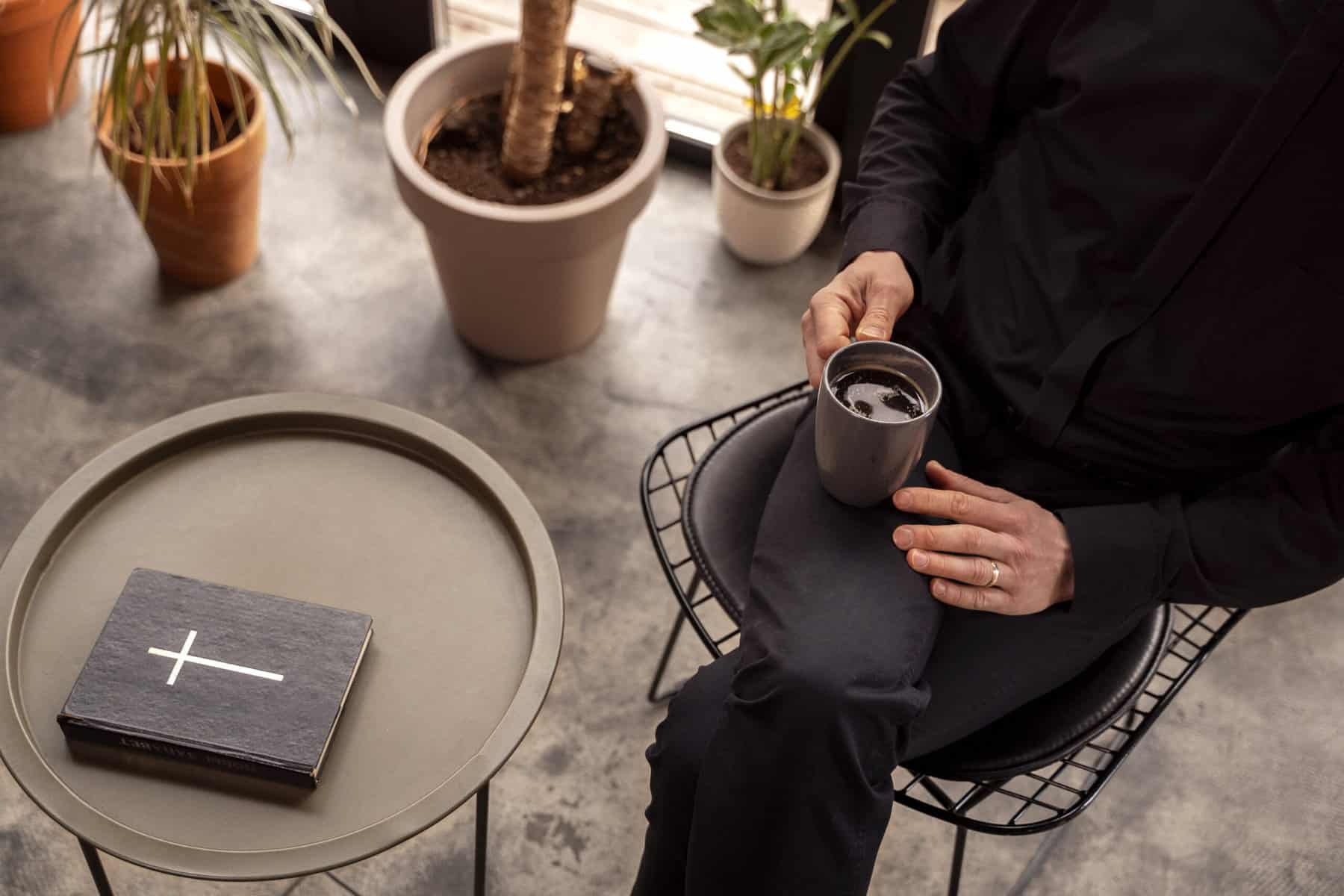 A person dressed in black sits with a cup of coffee in their lap, and a bible sits on a small table beside them