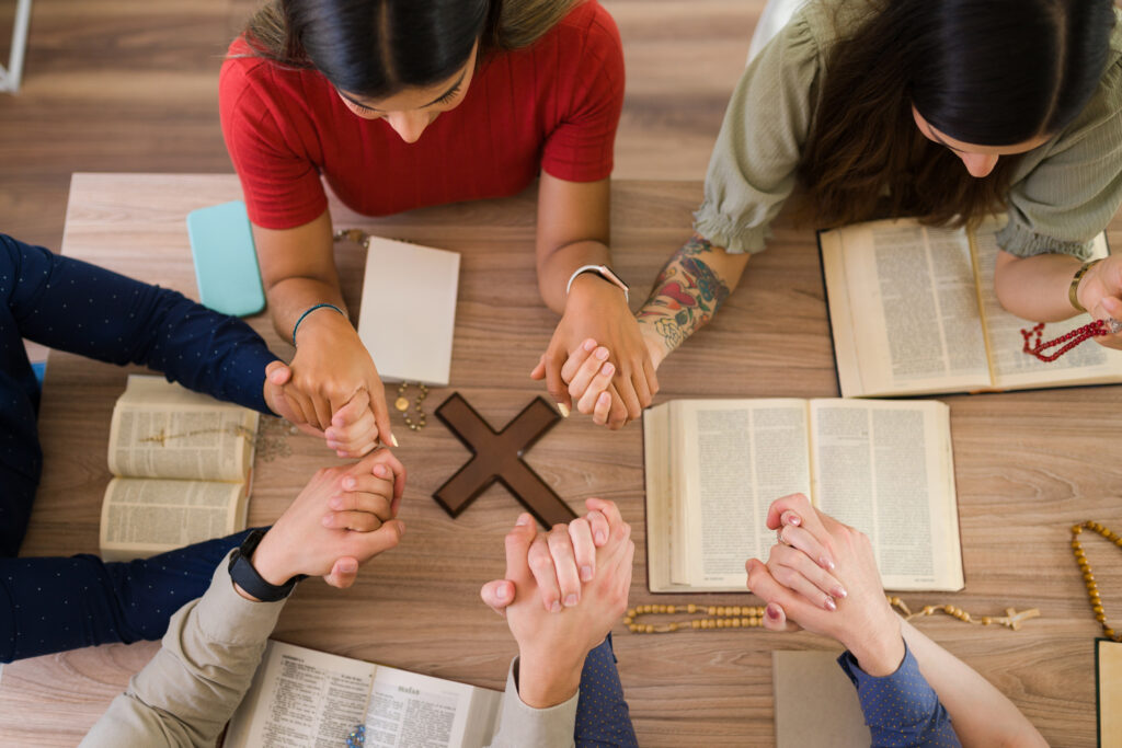 Five people are gathered around a table holding hands as seen from directly above. On the table sits open bibles and a wooden cross.