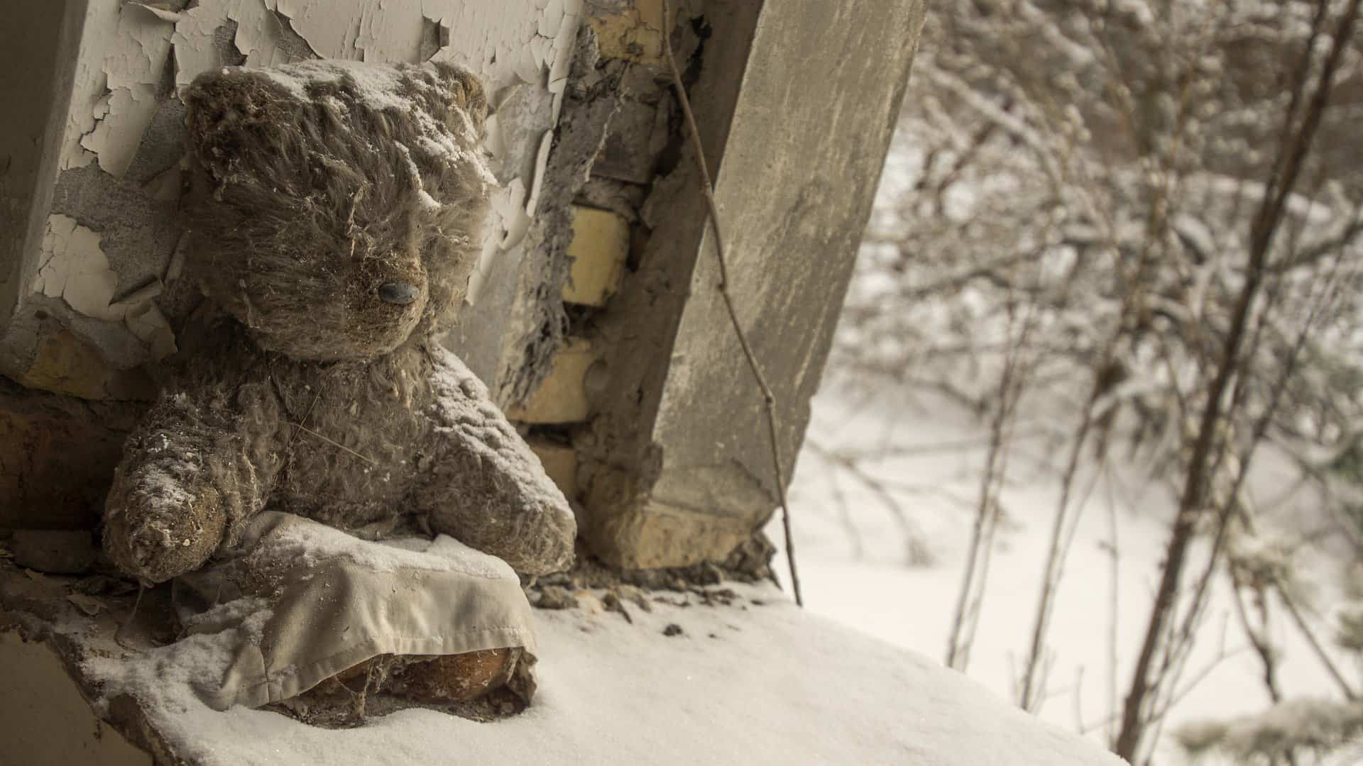 a sepia colored photo showing a well worn teddy bear covered in snow and sitting in a window frame of a derelict building