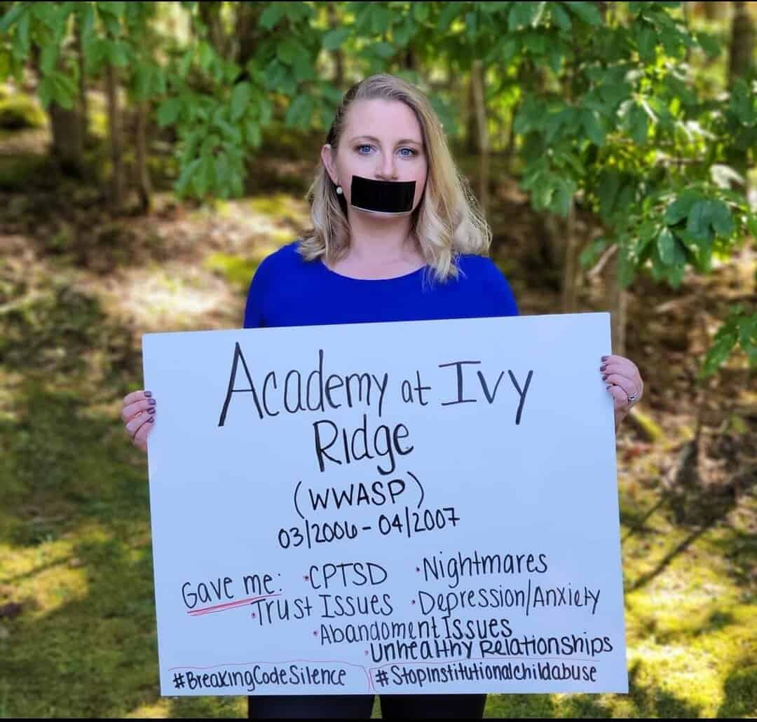 a thin young woman with blond hair holds a sign in front of her with problems she's faced due to attendance at Academy of Ivy Ridge. Her mouth is covered with black tape.