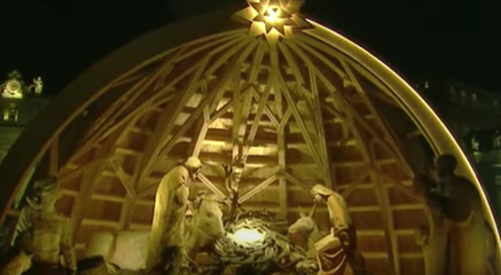 A nativity scene made of light colored wood sits under a dome of the same wood that is lit from the top by a soft light.