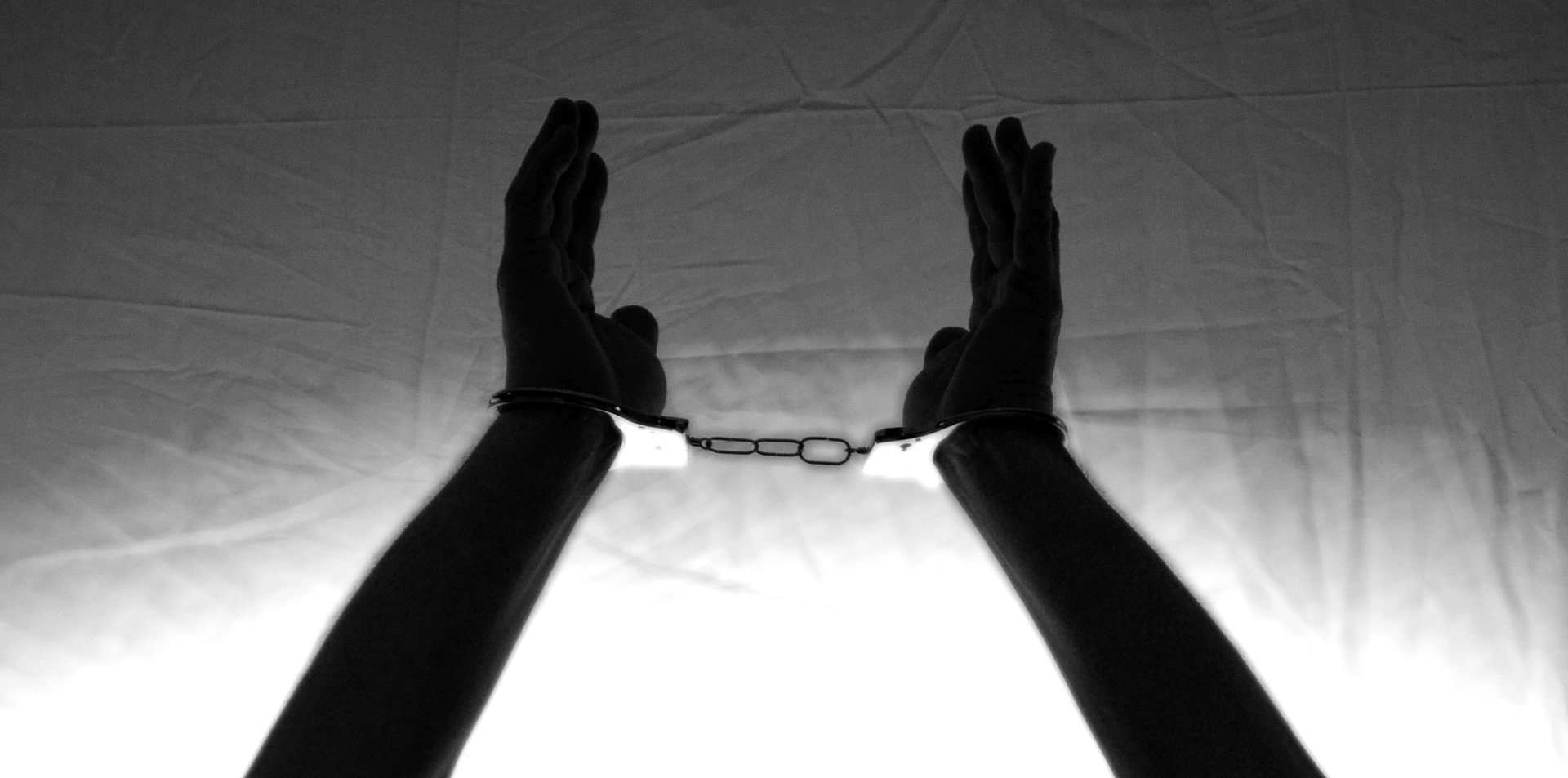 A person's hands and forearms are shown in black and white, lifted high. The wrist's are handcuffed.