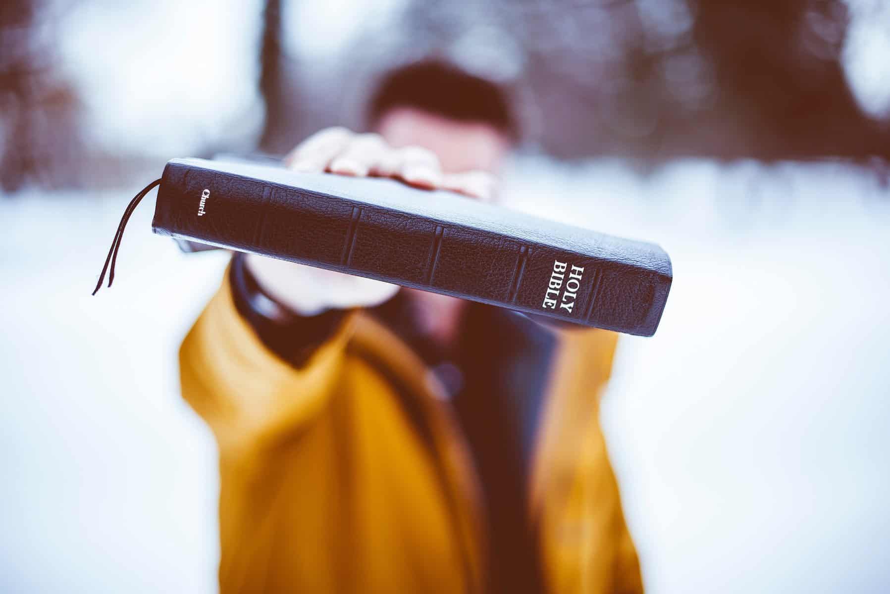 An out of focus man stands in the snow holding a black bible with the spine thrust toward the camera.