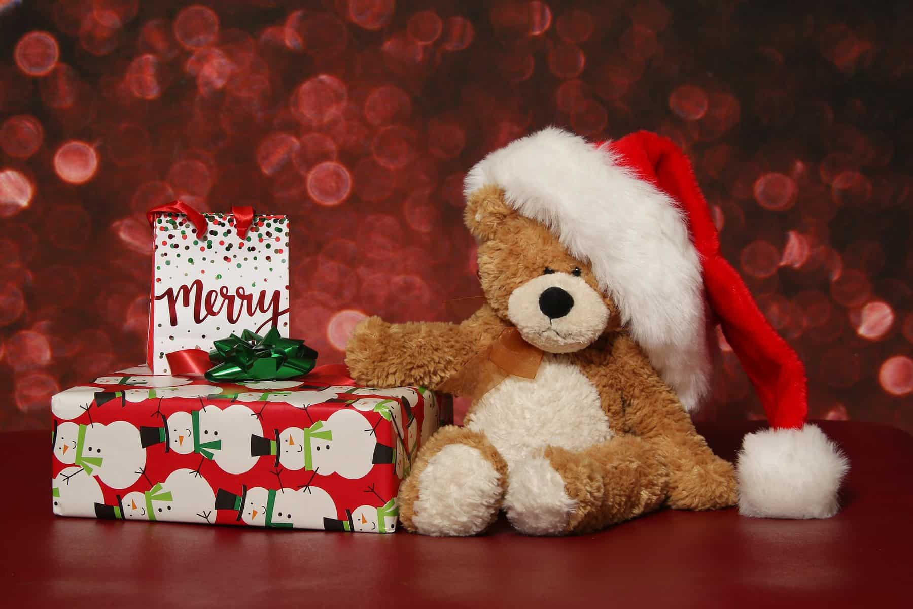 A brown teddy bear wearing a Santa hat sits next to a wrapped present with a Christmas card on it.