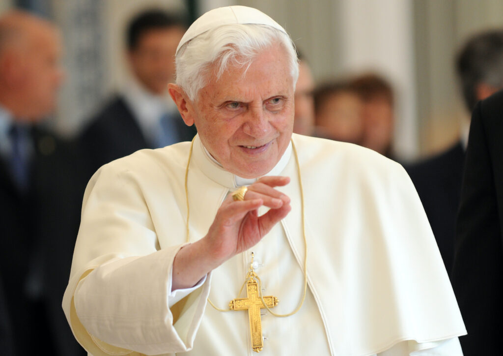 Pope Emeritus Benedict XVI is shown in off white Pope robes and gesturing to a crowd.