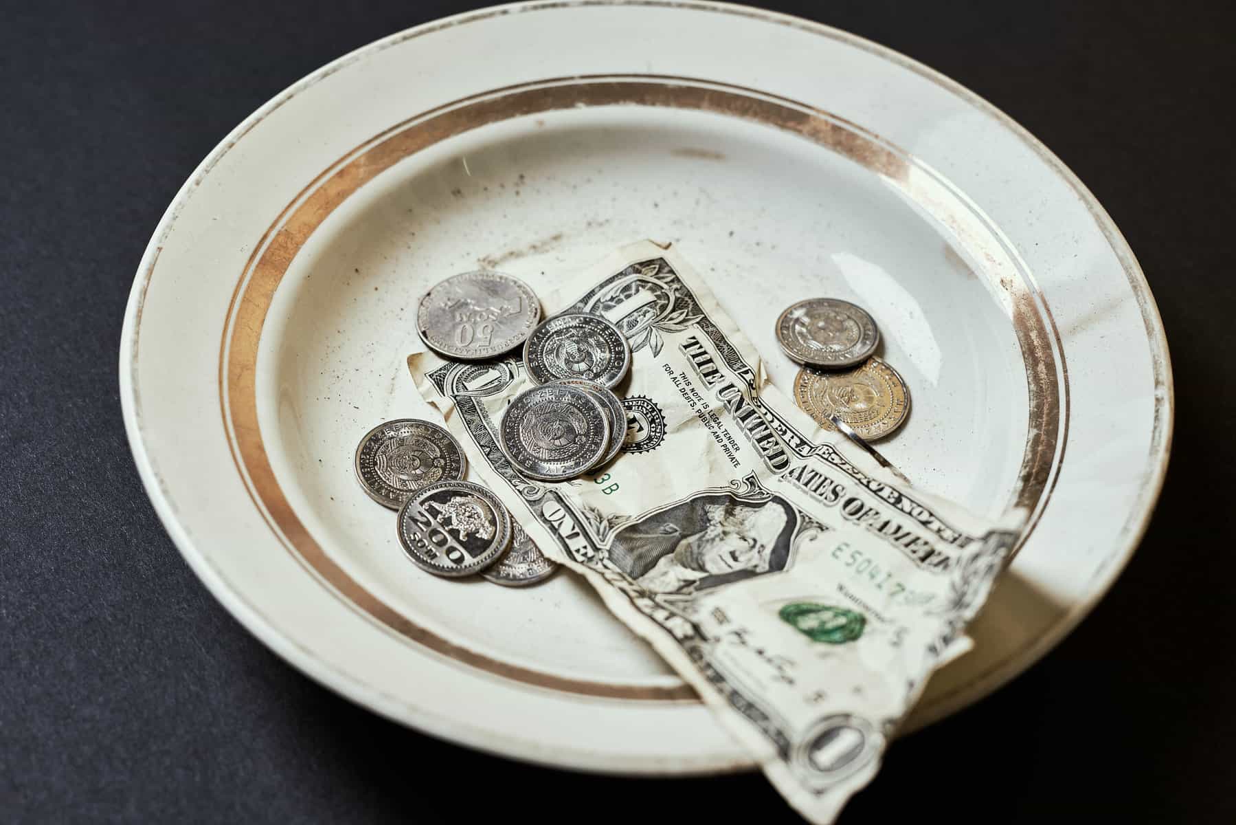An old off white plate with worn gold around the edges holds donated money in coins and a bill.