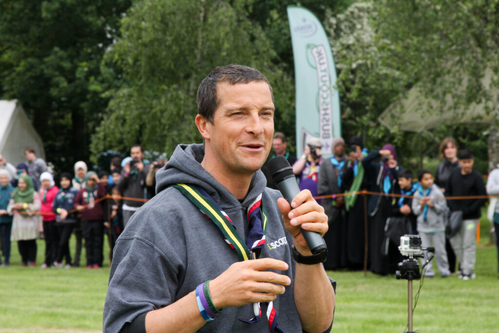 Bear Grylls is shown in casual clothes holding a microphone and speaking at an outdoor event.