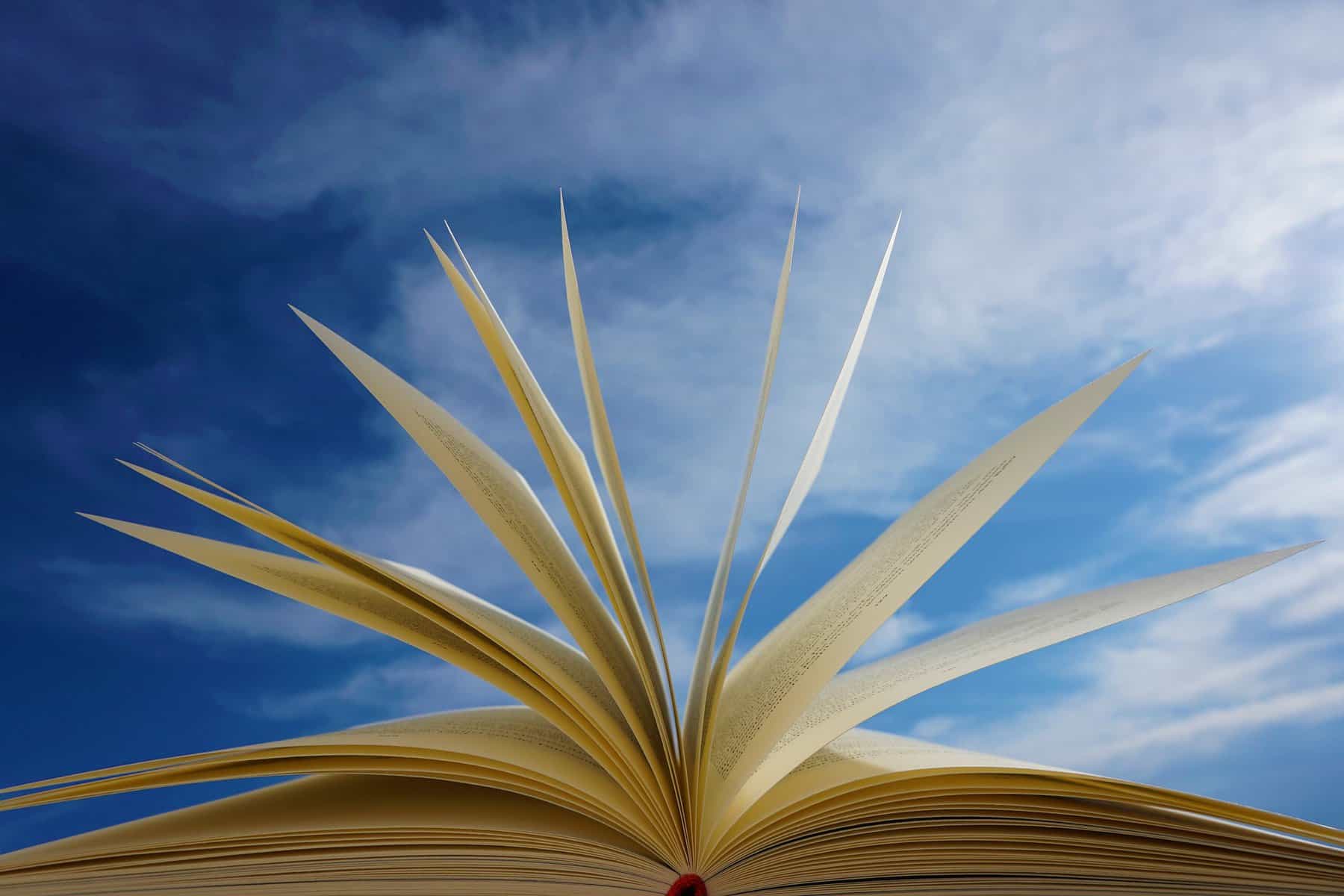 An open book laying on a flat surface is shown from the bottom with pages at various angles. A blue and cloudy sky is the backdrop.