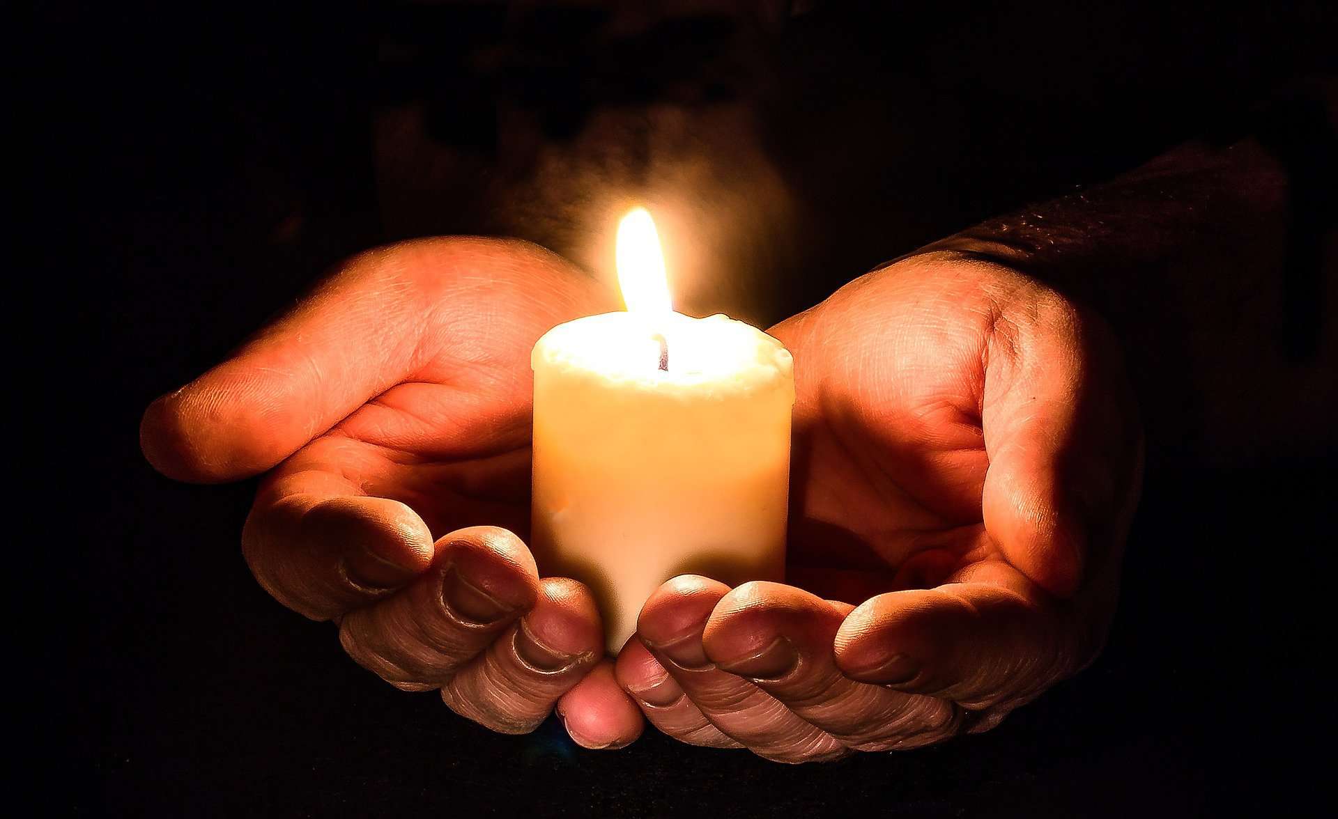 Hands shown close up hold a small, lit, votive candle.