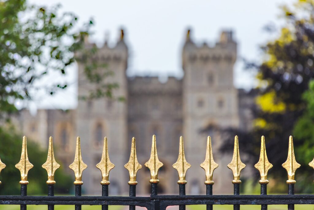 Image shows gold finials on black wrought iron fence with a castle like building with two towers blurred in the distance.