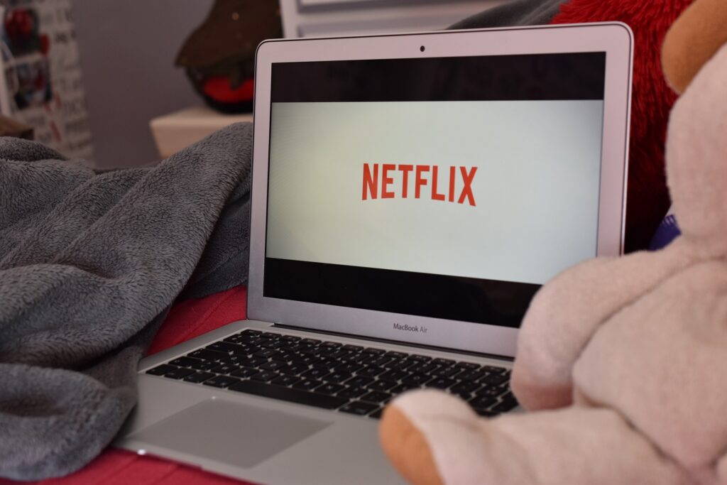A laptop sits in what looks like a home environment with a stuffed bear next to it and the Netflix logo on the screen.