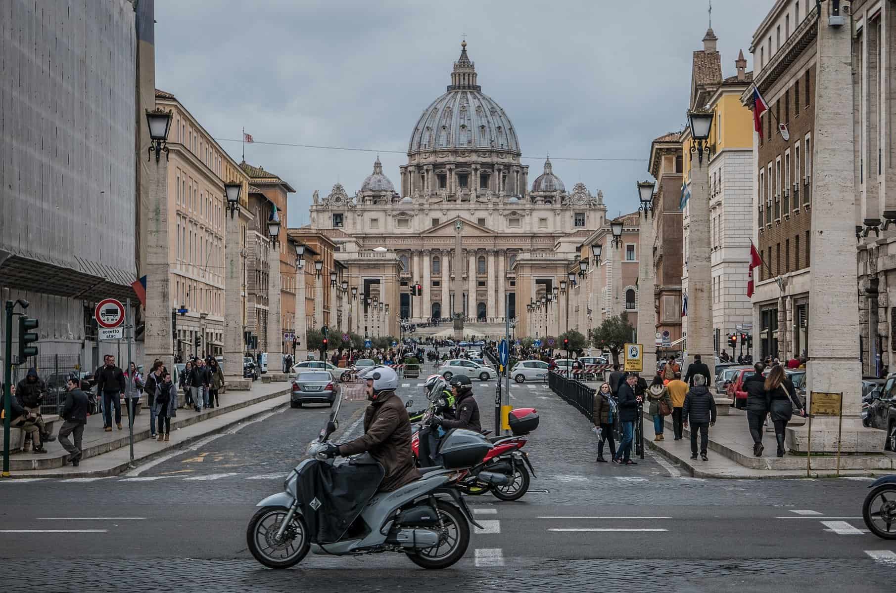 A motorbike and cars are shown in the foreground on a street with the Vatican dome in the distance.