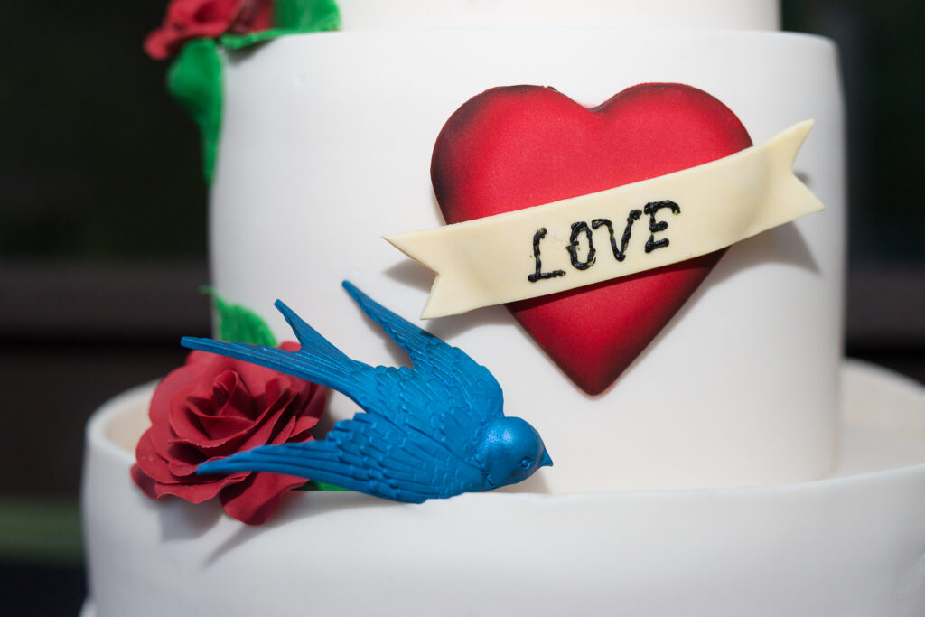 A white cake is shown with a red heart on which is written "love" with a blue bird is shown below.